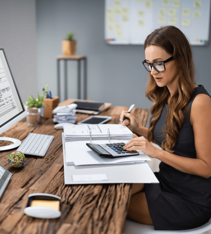 Bookkeeper working at desk with calculator, computer, and financial documents.