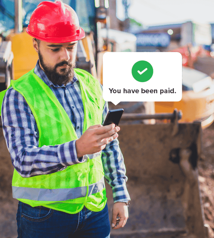 Construction worker in a red hard hat and neon vest checking his phone at a job site, with an on-screen message that says, “You have been paid.