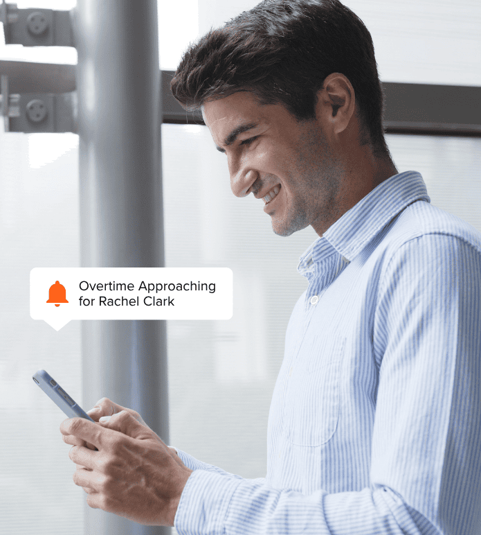 A manager smiles while looking at his smartphone, which displays a notification alert that reads: Overtime Approaching for Rachel Clark.