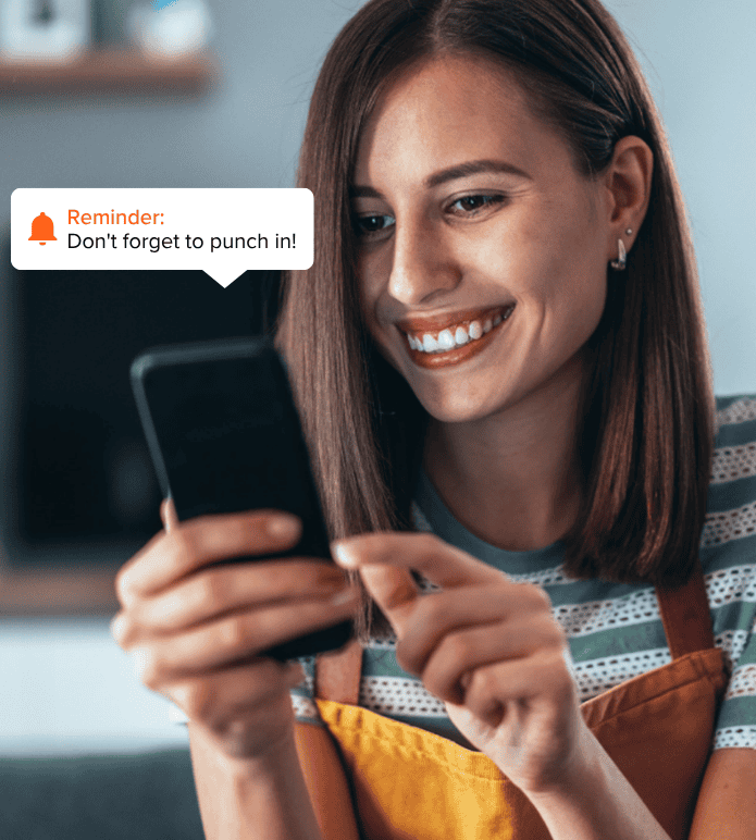 Smiling woman in an apron looking at her phone, with a reminder notification saying, “Don’t forget to punch in!